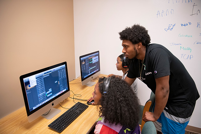 Camp youth counselor help students during STEAM Camp in front of computers.